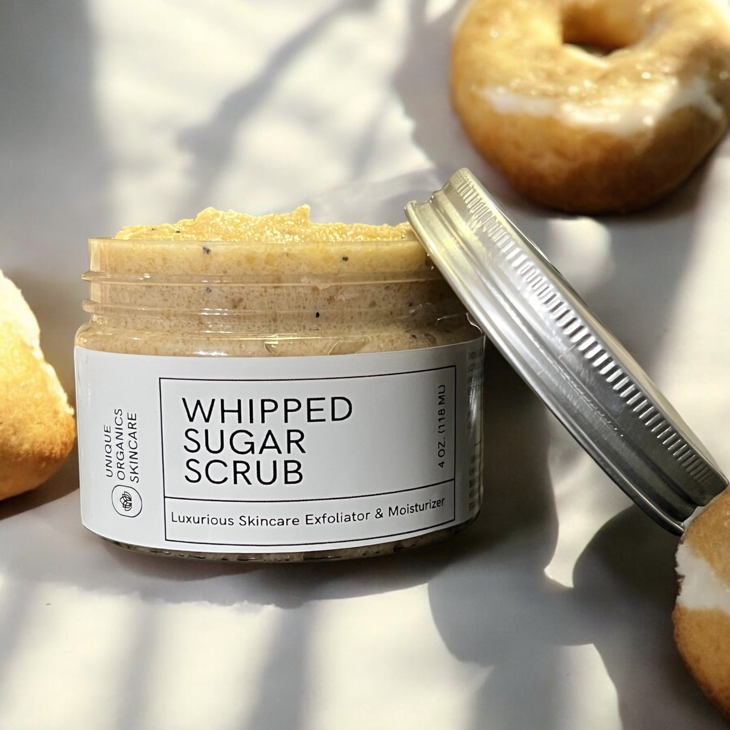Jar of whipped sugar scrub with a lid off, surrounded by bagels on a light surface.